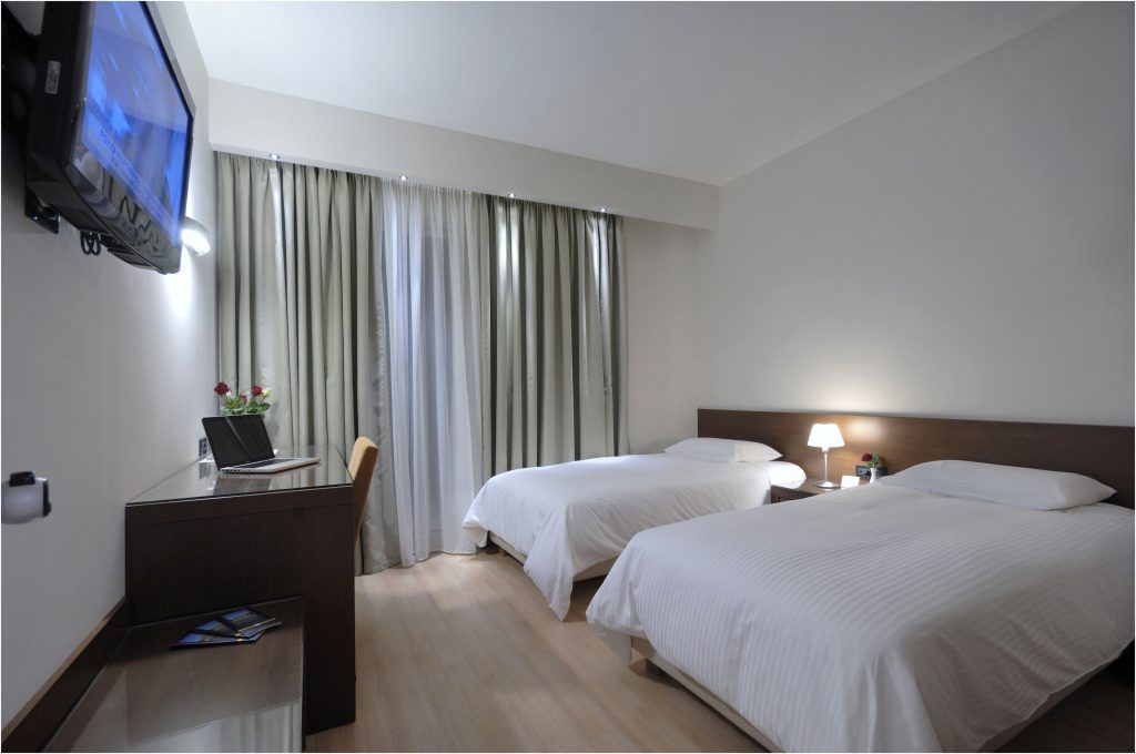 Two single beds with white linens, flat-screen TV, and desk with laptop and flowers.